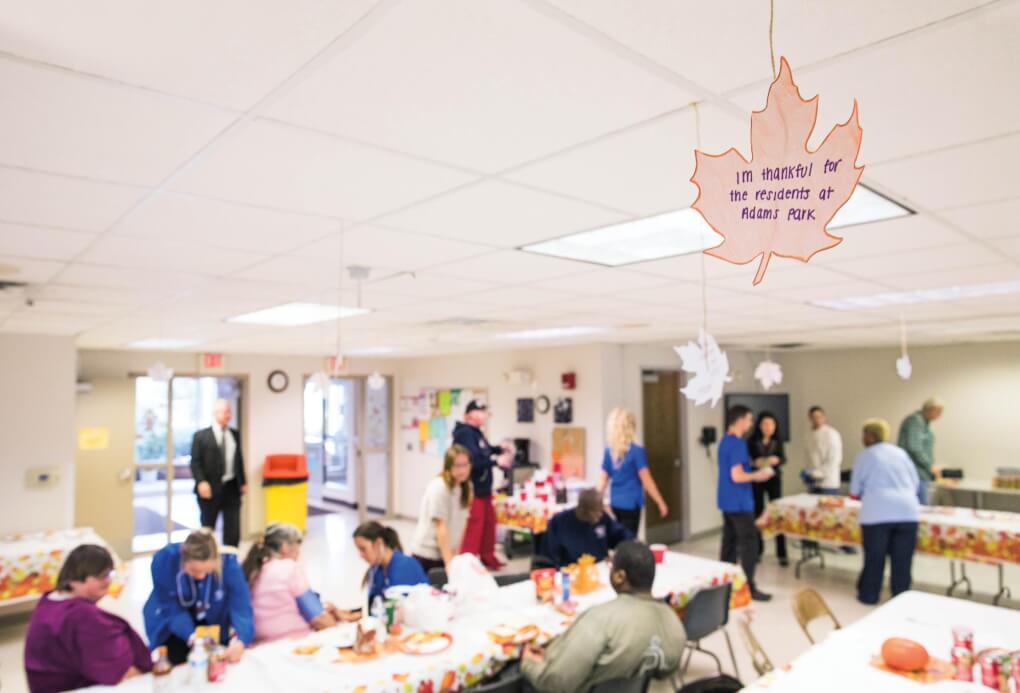 Cut out leaf with words 'I'm thankful for the residents at Adams Park' written on it. Room set up for thanksgiving with people in background.